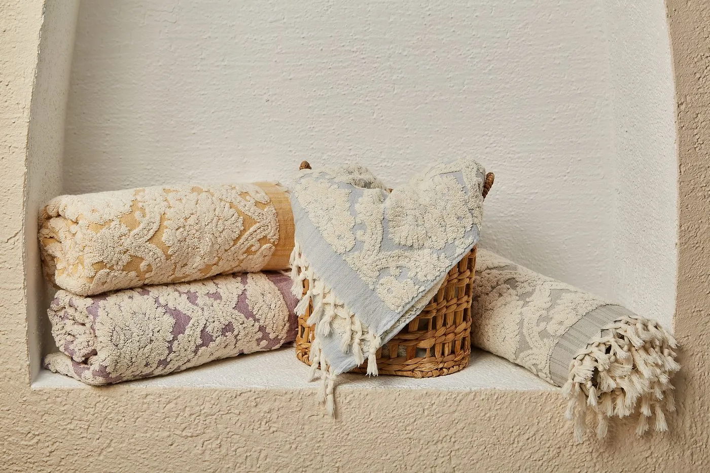 Rolls of damask-woven Turkish bath towels in gold, lilac and blue stacked in a whitewashed niche